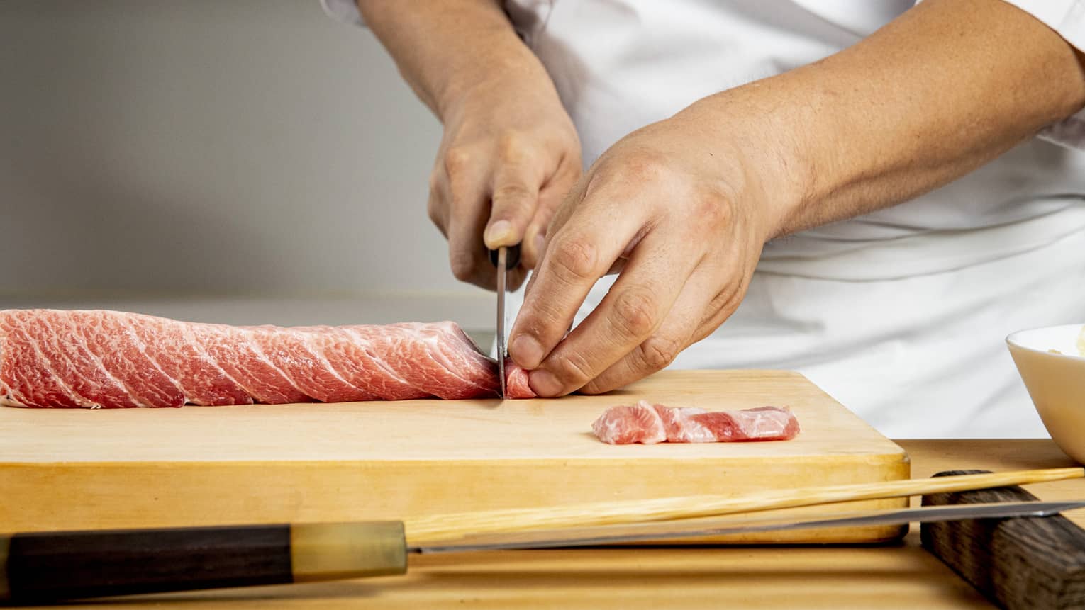 Chef's hands use knife to thinly slice meat on wooden board, preparing it for sushi