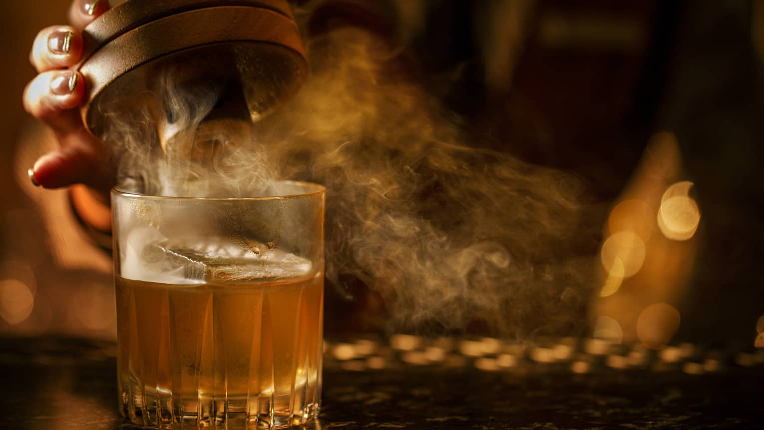 Hand lifts the lid of a smoked Old-Fashioned cocktail in a dimly lit setting