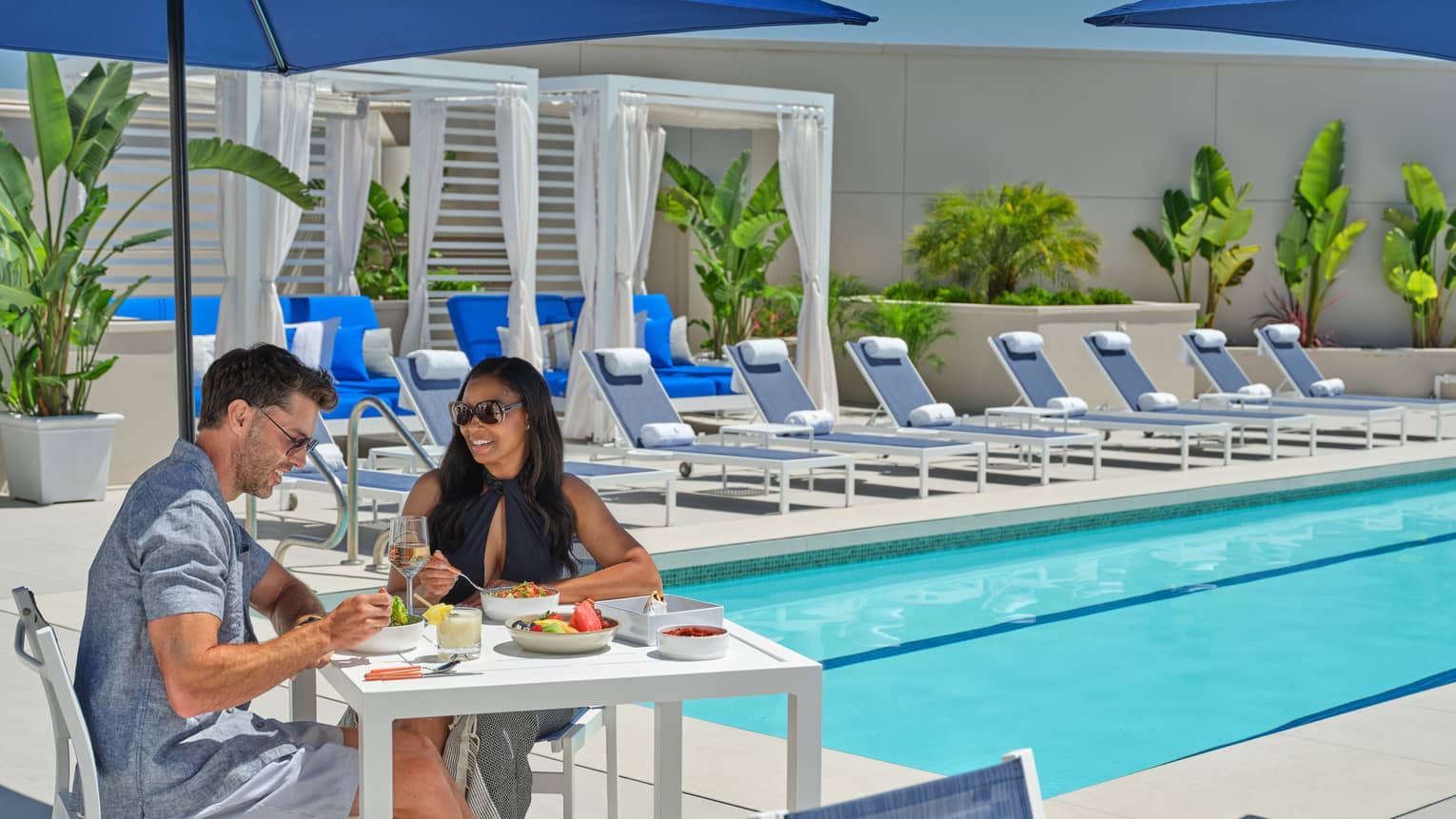 Two guests sitting at a table under a blue umbrella by the pool.