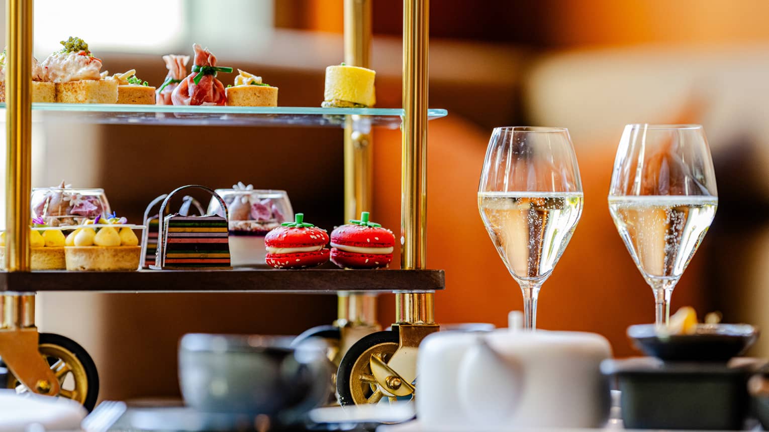 A tiered serving tray on small wheels holds colourful small bites and is sitting on a table next two two stemmed glasses holding a light-hued liquid, a teapot, and a teacup and saucer.