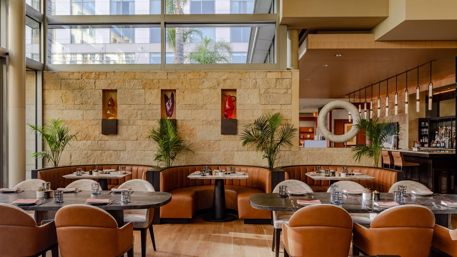 A restaurant with windows near the ceiling, leather chairs and plants beside the tables