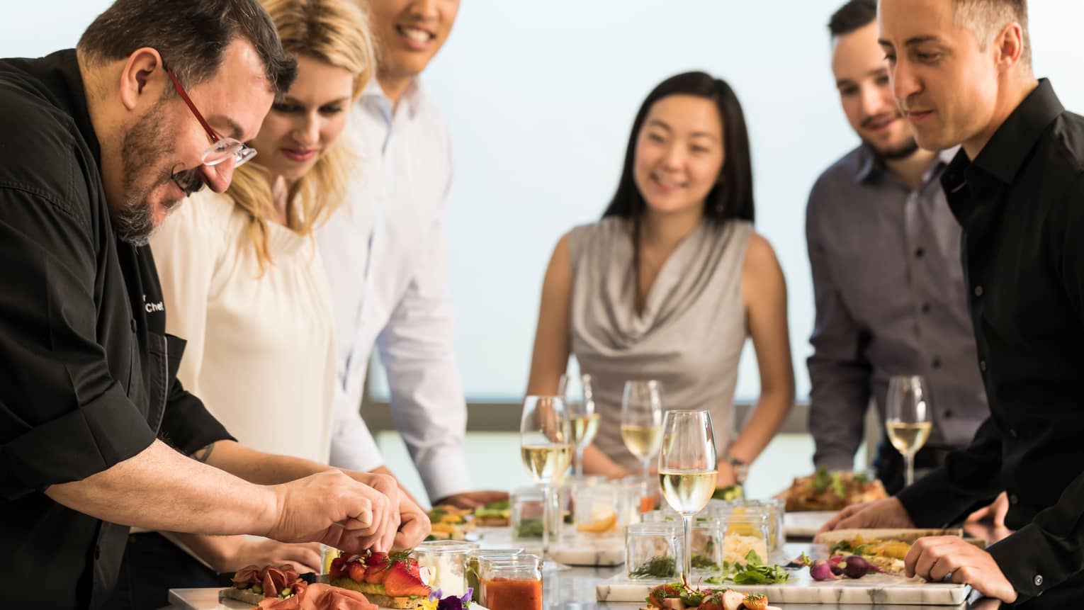Chef assembles food at counter with smiling group of friends, white wine in glasses