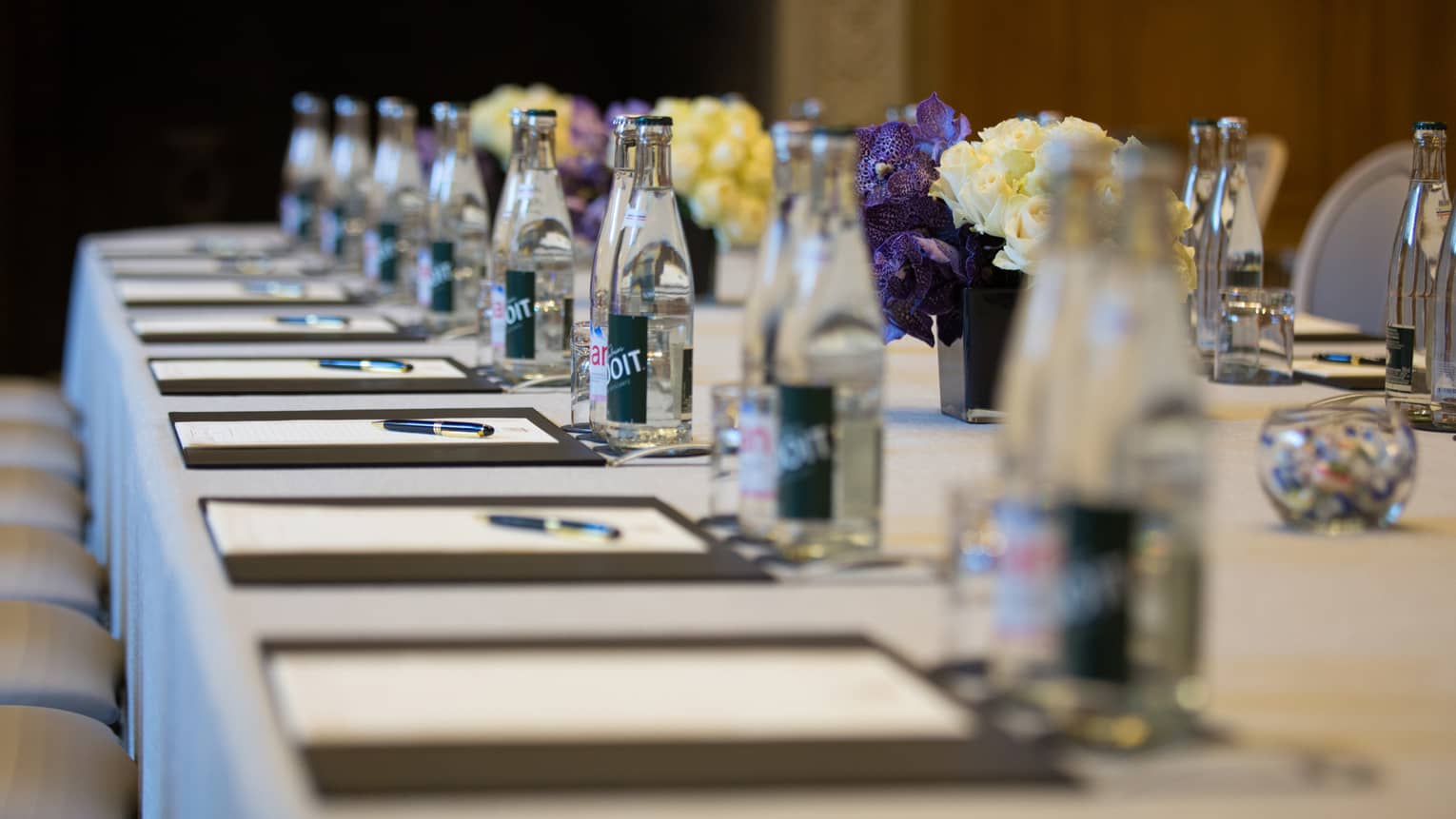 Row of meeting agendas, glass bottles, small vases with flowers on boardroom table