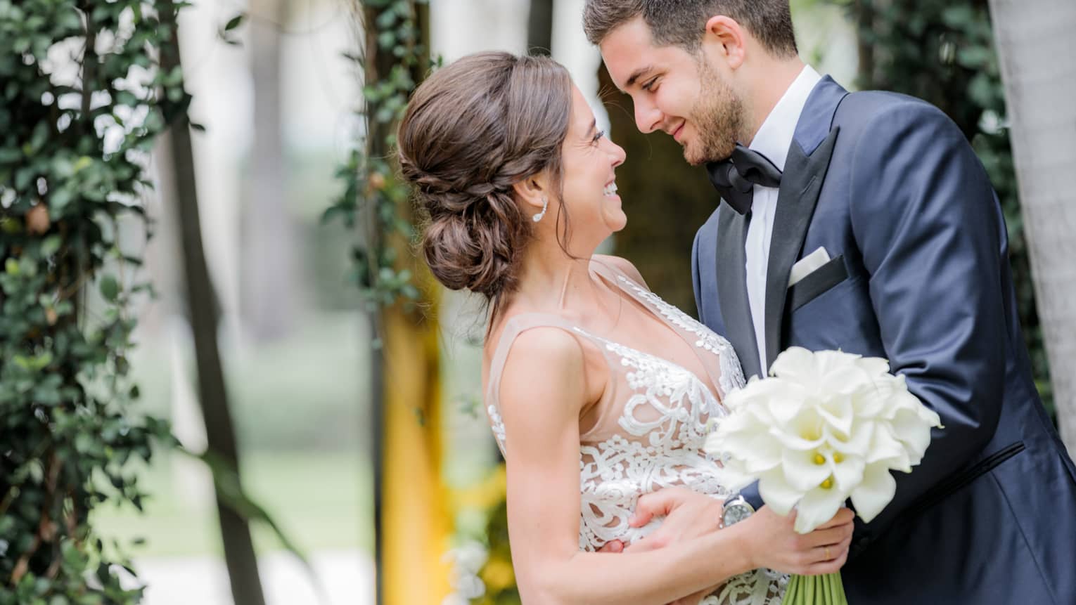 Smiling groom and bride with white wedding dress, bouquet embrace
