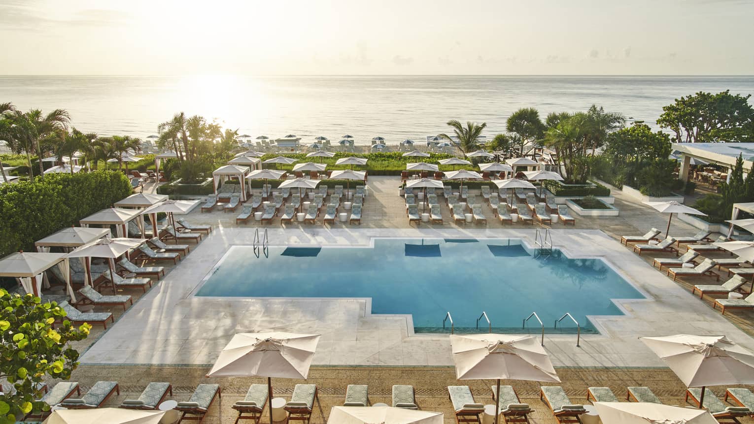 A pool surrounded by lounge chairs and large umbrellas with the ocean in the background.