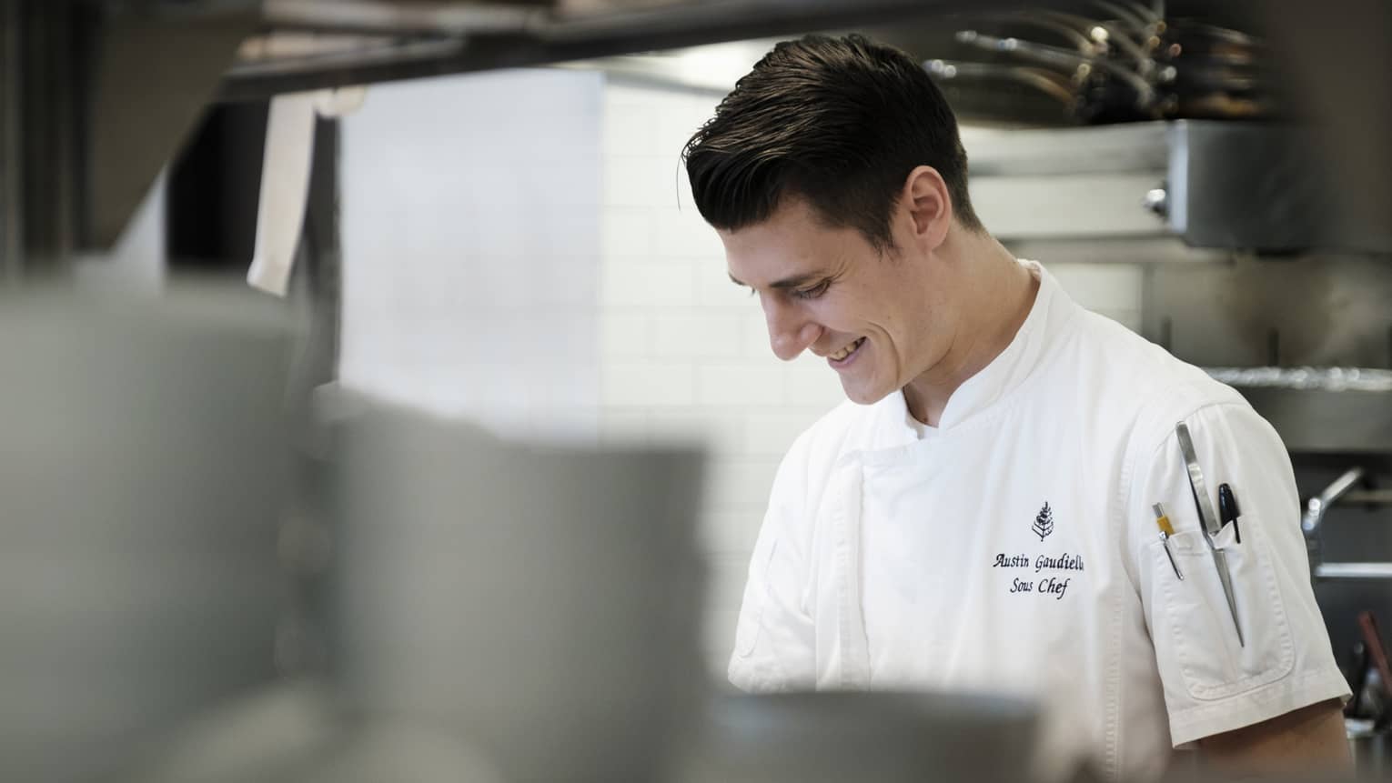 Person wearing a chef?s coat and smiling while standing in the kitchen.