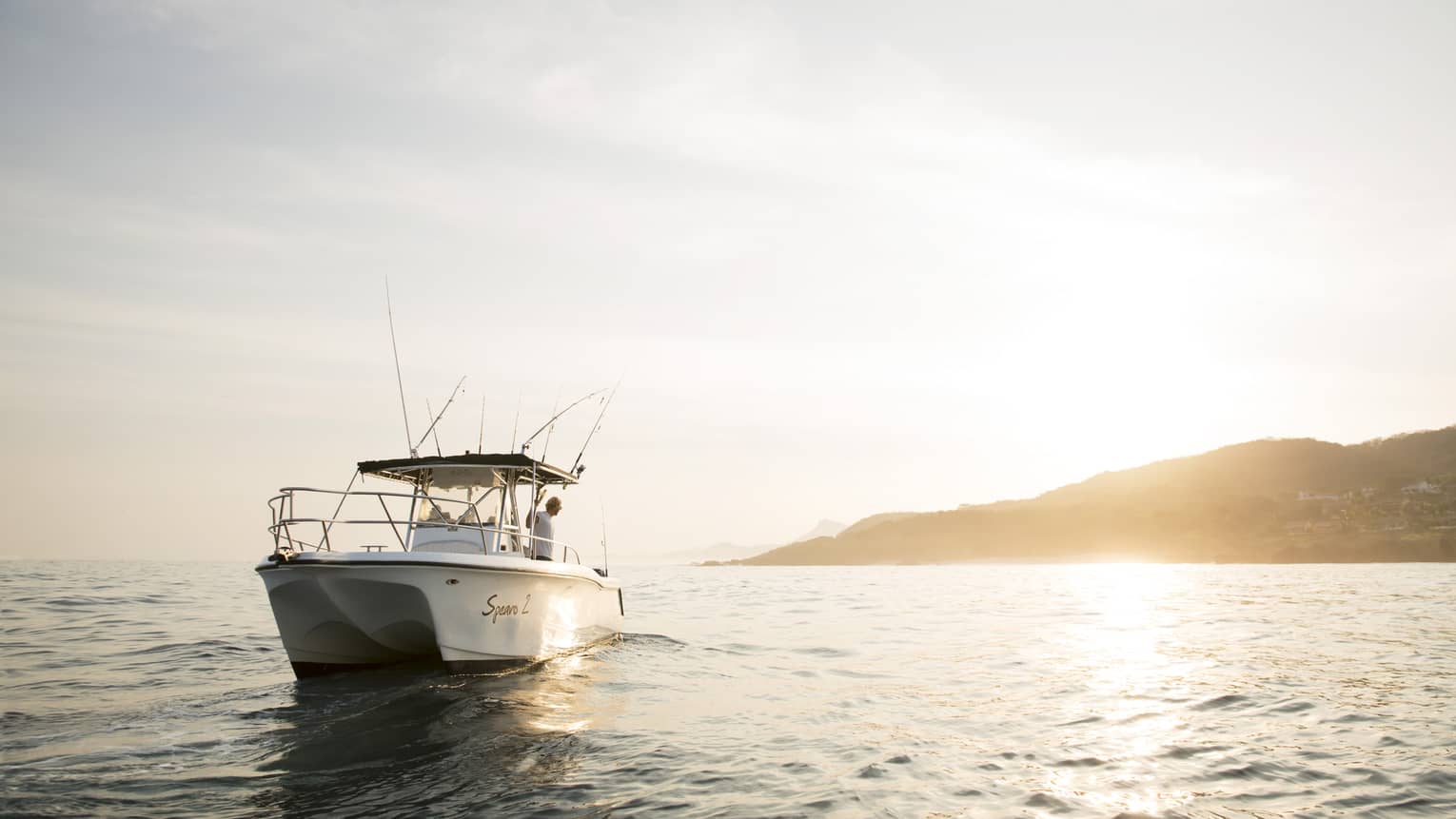 Front long view of a white boat bobbing in gentle waves with a person onboard looking out at the sun reflecting on the water.