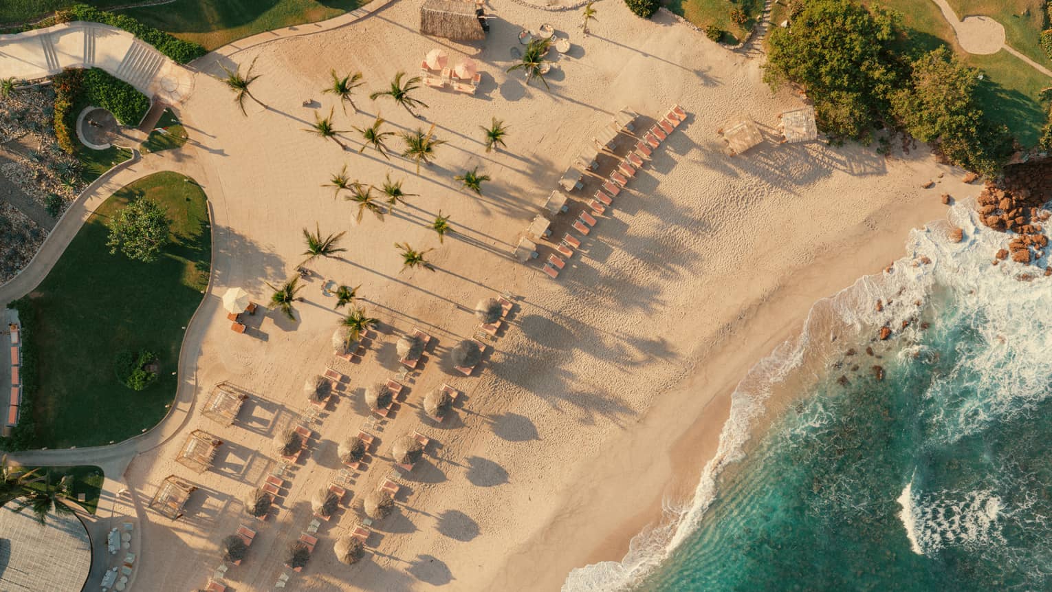 A bird's-eye view of a sandy beach next to turquoise waters. The beach is dotted with rows of lounge chairs and umbrellas and palm trees.