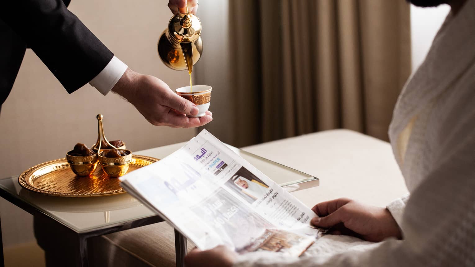 Man wearing bathrobe sits on chaise with newspaper and is served coffee