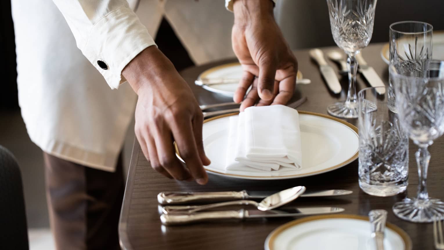 The hands of a server wearing a long-sleeved button down shirt adjusts a gold-rimmed white plate on a table set with various plates, silver cutlery and crystal glassware