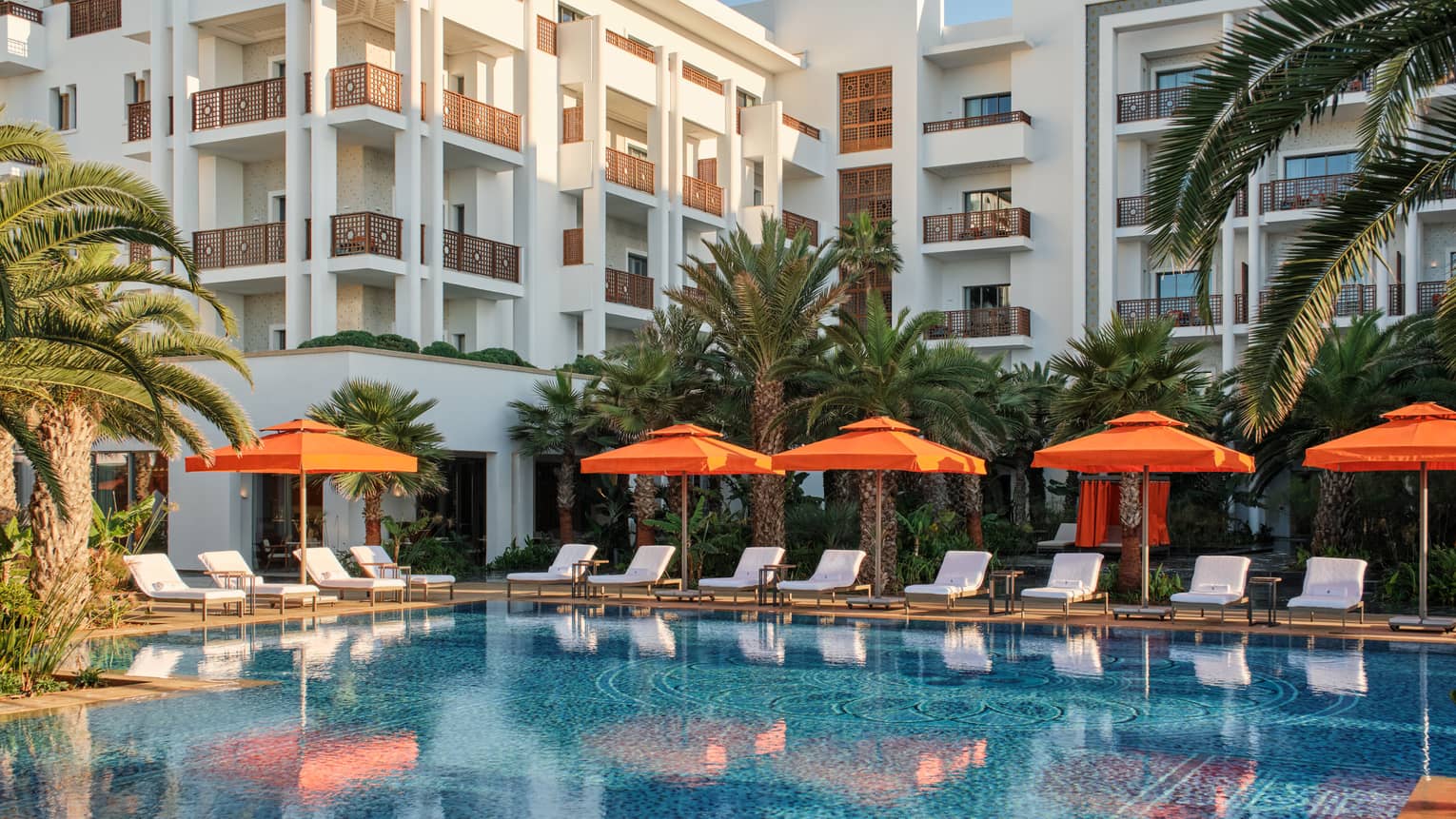 Pool lined with orange umbrellas by white lounge chairs outside of white building and surrounded with palm trees