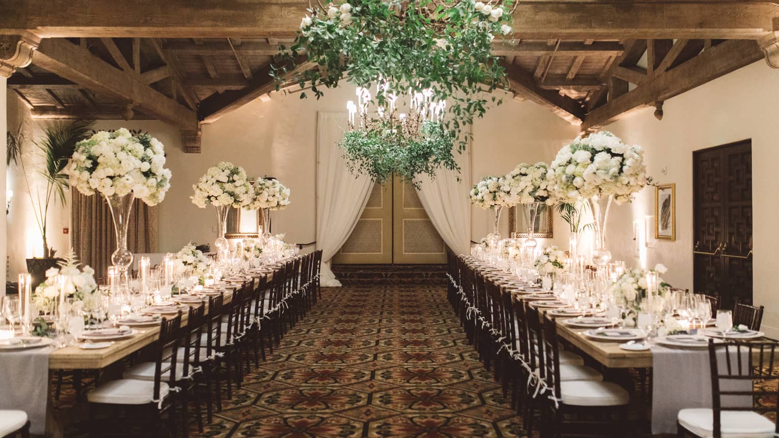 Two long wedding dining tables under chandelier with vines, wood beam ceiling