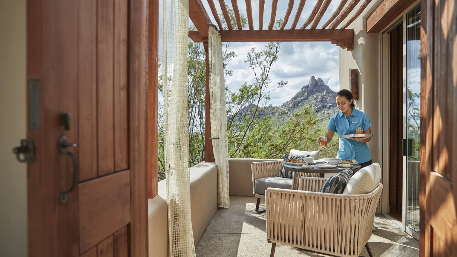 Hotel staff placing food on a hotel room's outdoor terrace, two arm chairs, wooden pergola