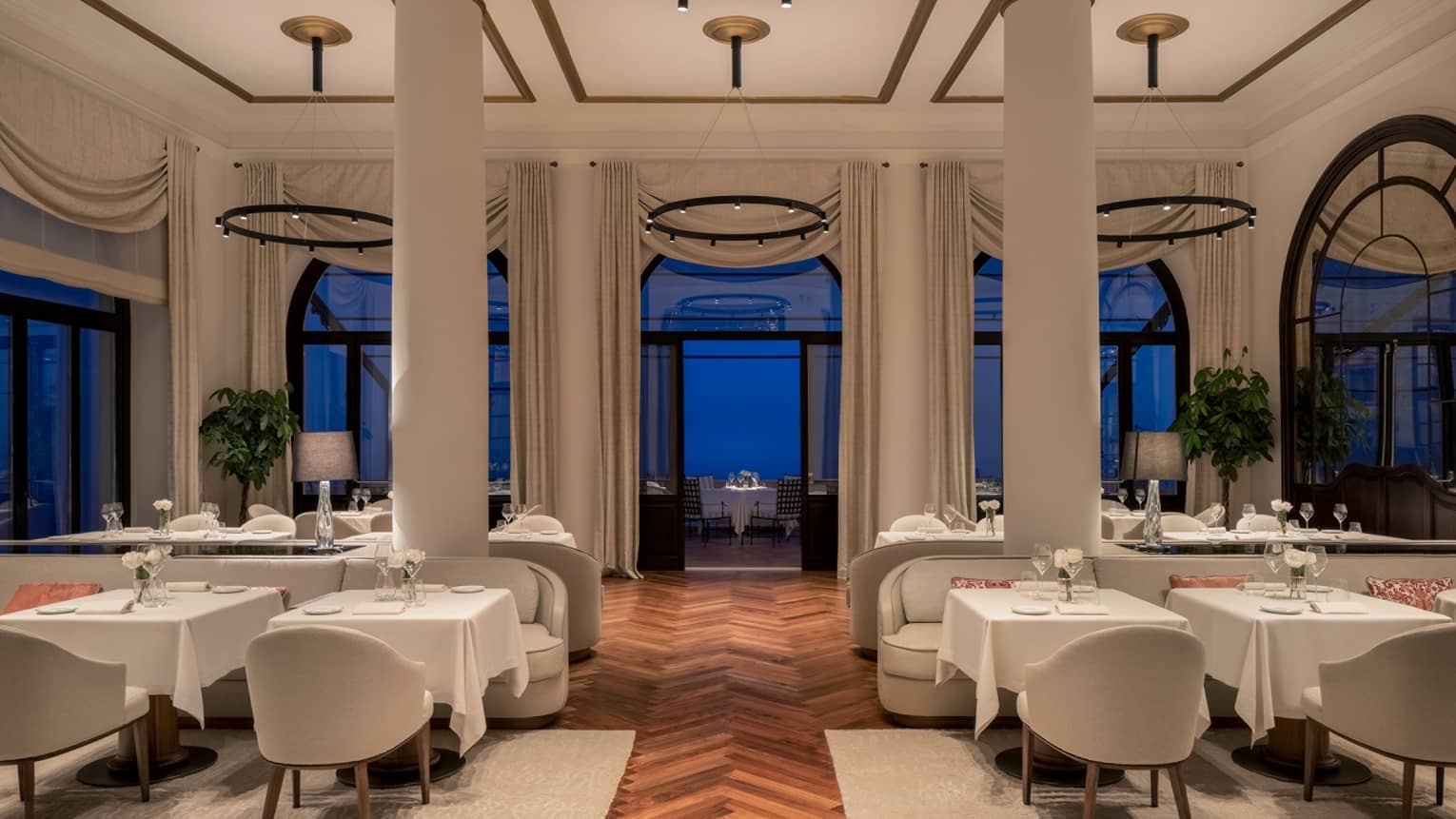 A restaurant with columns, large windows and white cloth tables.