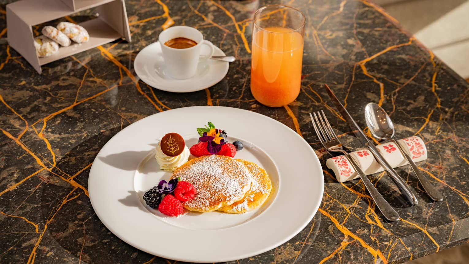 Two pancakes topped with powdered sugar and garnished with mixed berries and butter beside flatware, cup of espresso and glass of juice