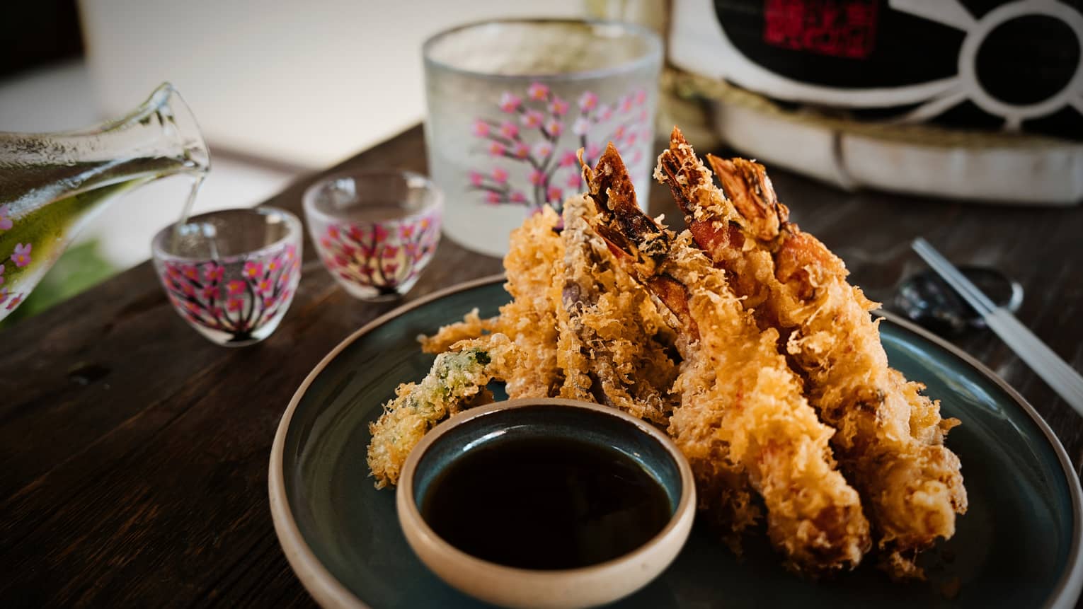 A plate of Shrimp Tempura with sauce on a dark plate with someone pouring a beverage into a small glass in the background.