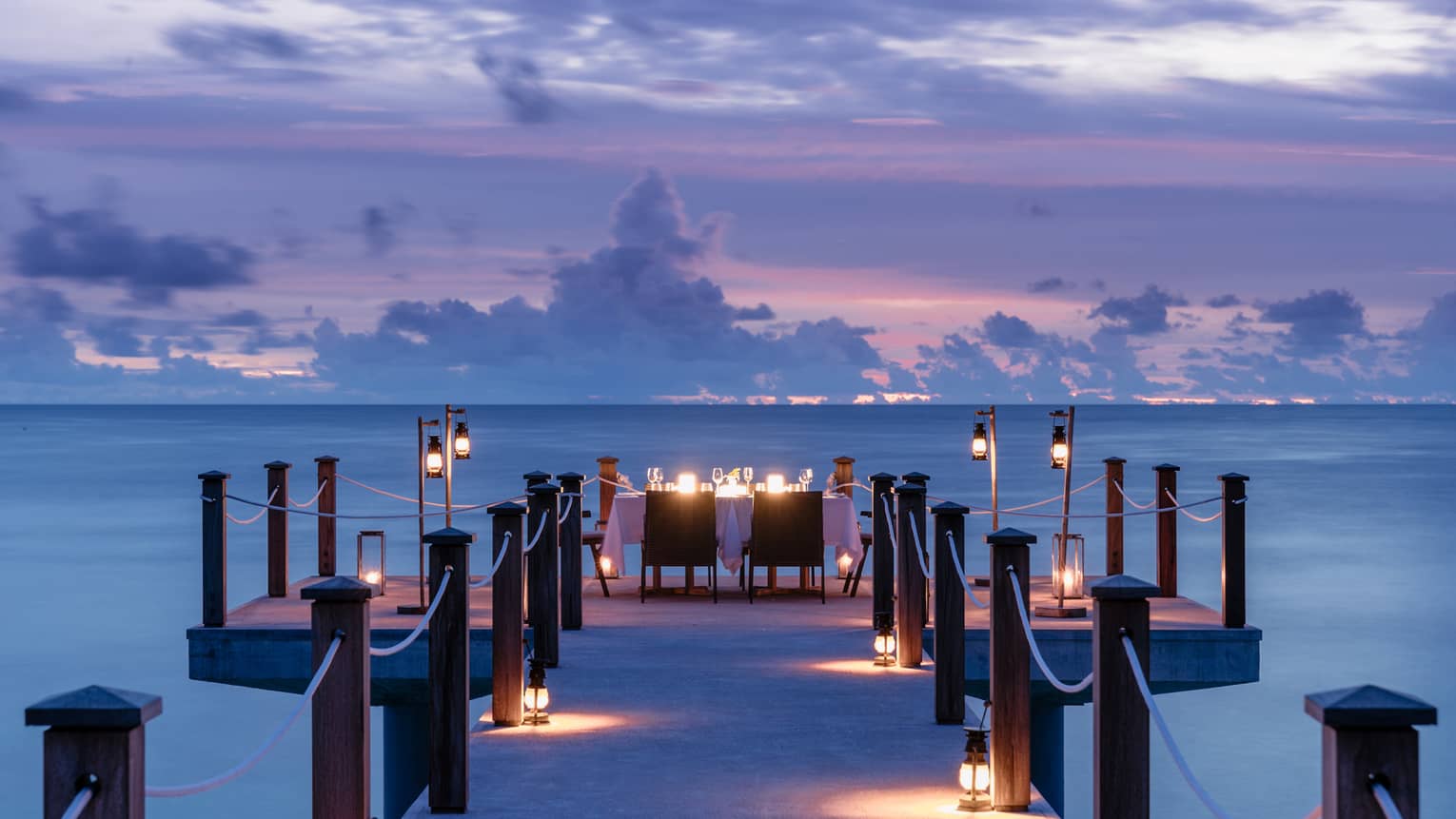 Romantic dinner table at end of pier overlooking ocean at dusk, surrounded by lanterns