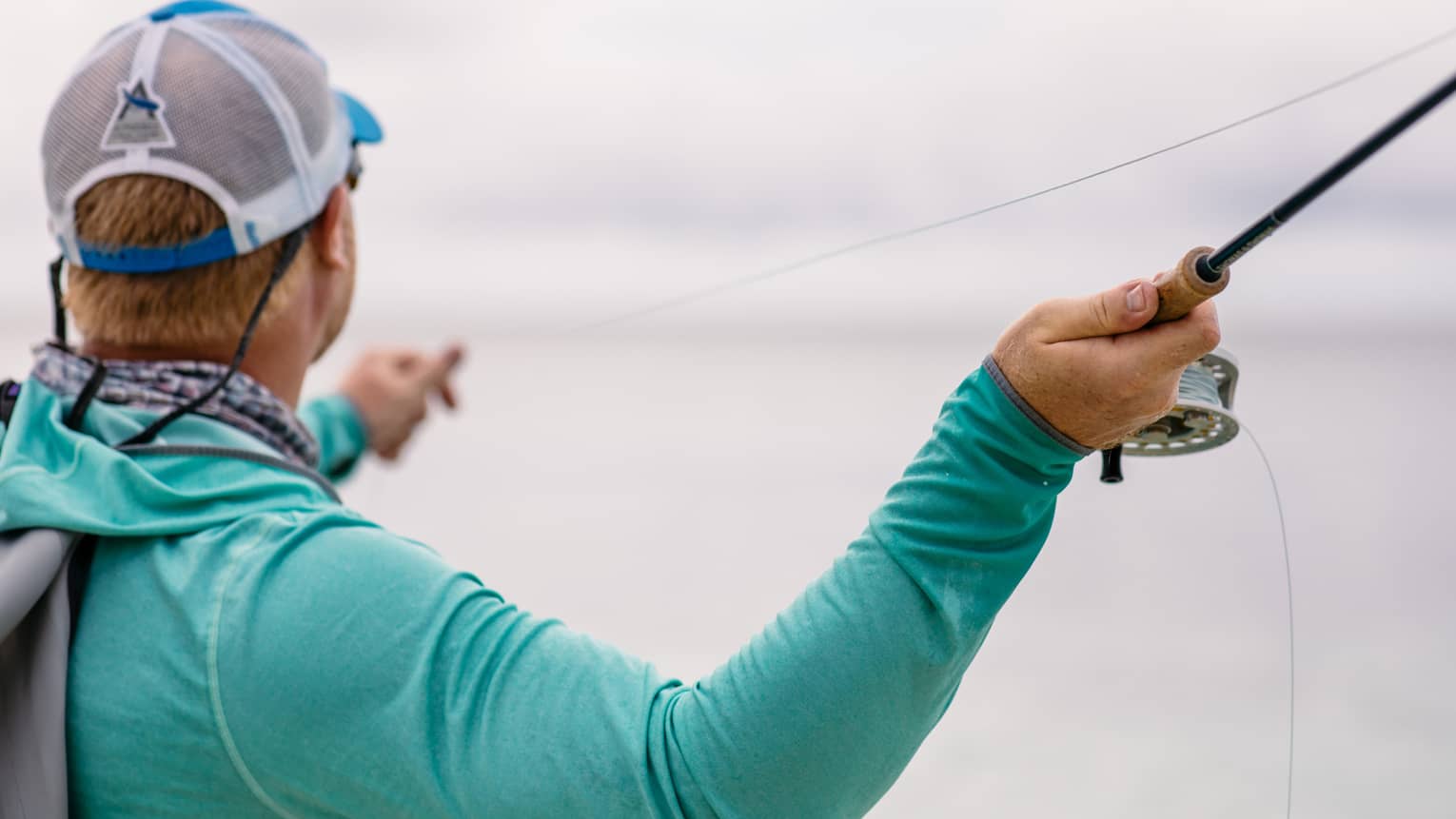 A fisherman facing away holds his fishing line in his left hand and draws back his rod with his right, readying to cast.