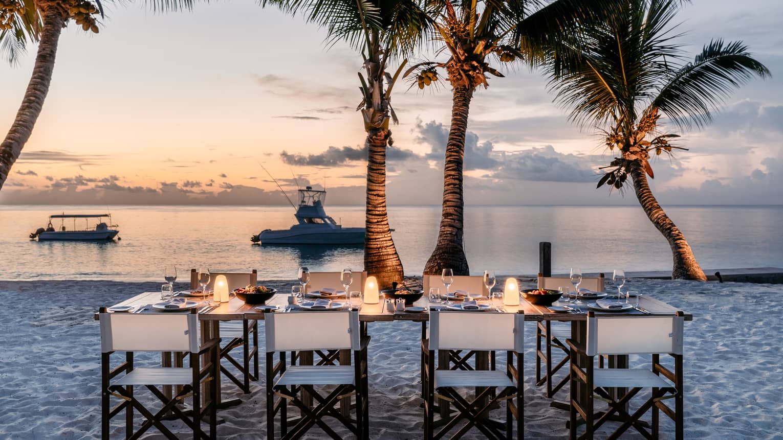 Eight director's chairs around rectangular candlelit table set on beach with palm trees at sunset