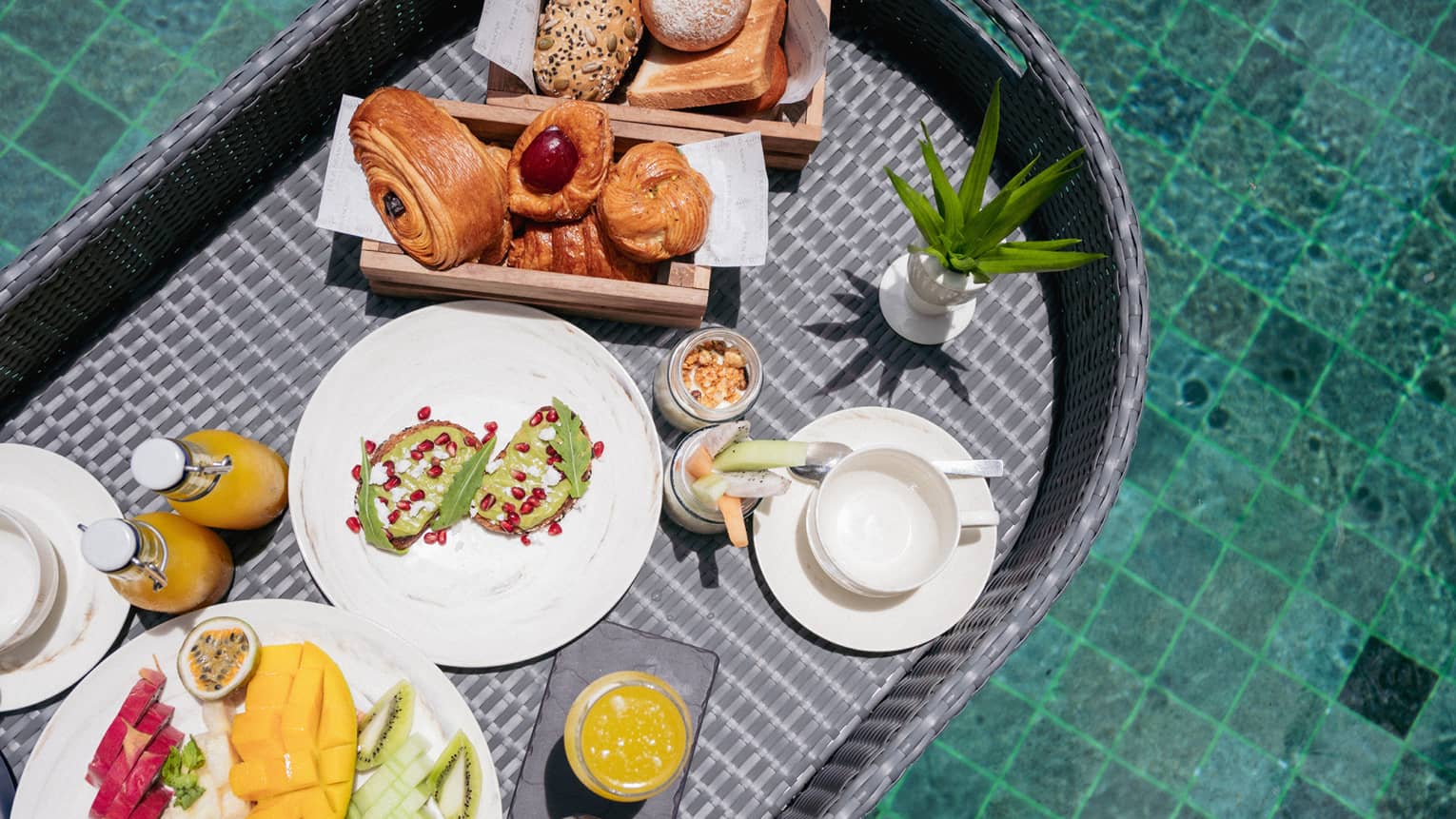 Close-up view from above of a wicker tray filled with fruit, pastries, juice and toast floating in a crystal-clear pool.