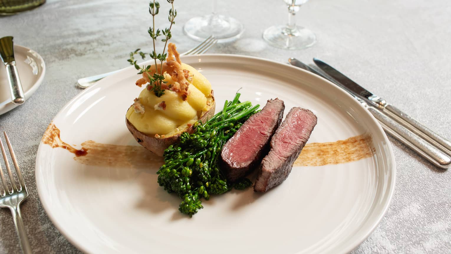 Round white plate of medium-rare steak, blanched broccolini and stuffed baked potato