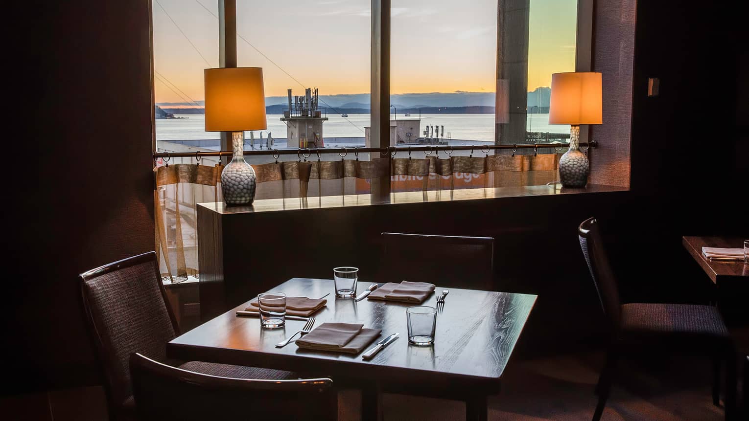 Dimly-lit dining table by two small lamps and window with sunset, ocean view 