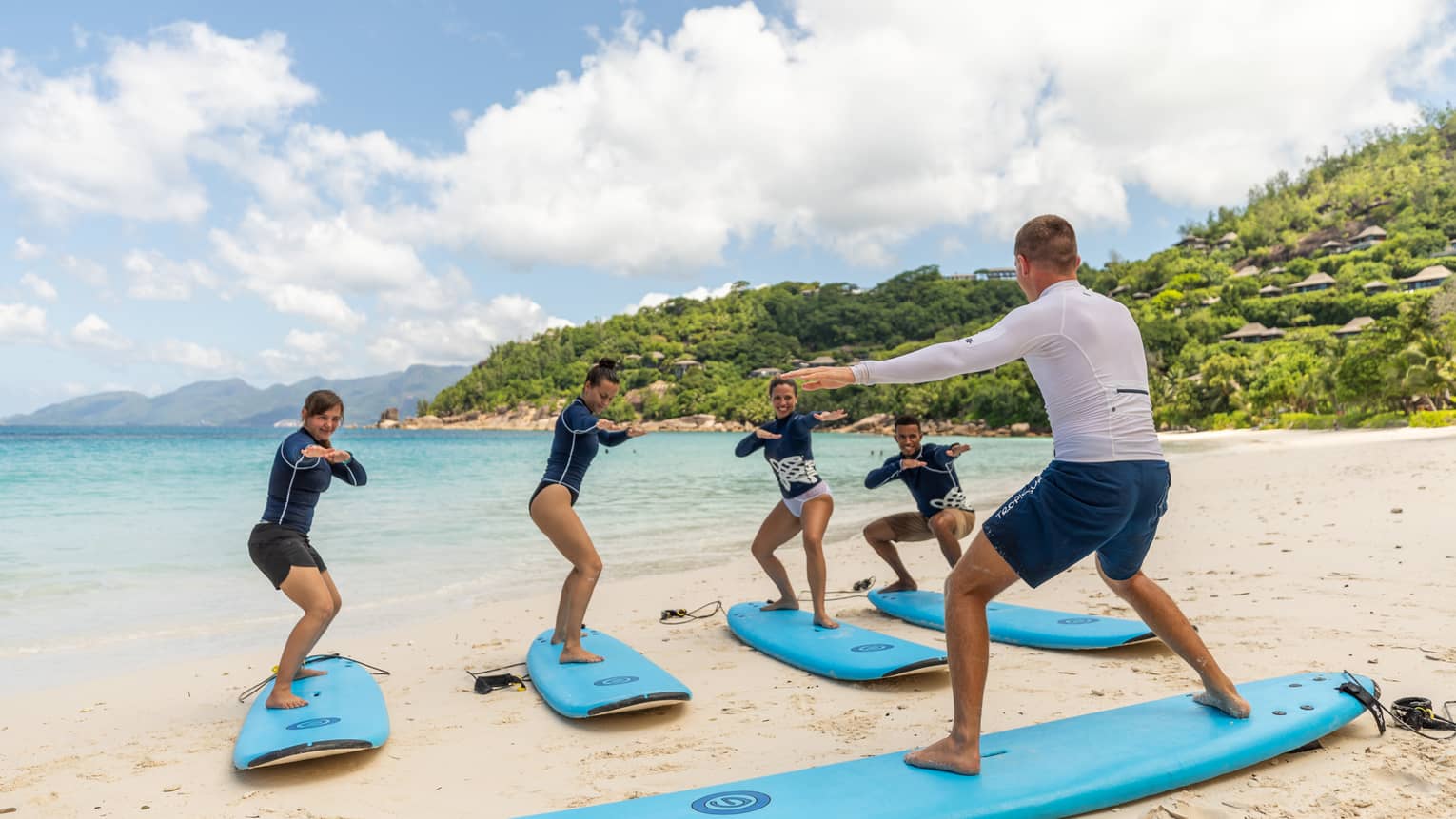 Surfing instructor balances on surfboard on white sand beach in front of row of surfers