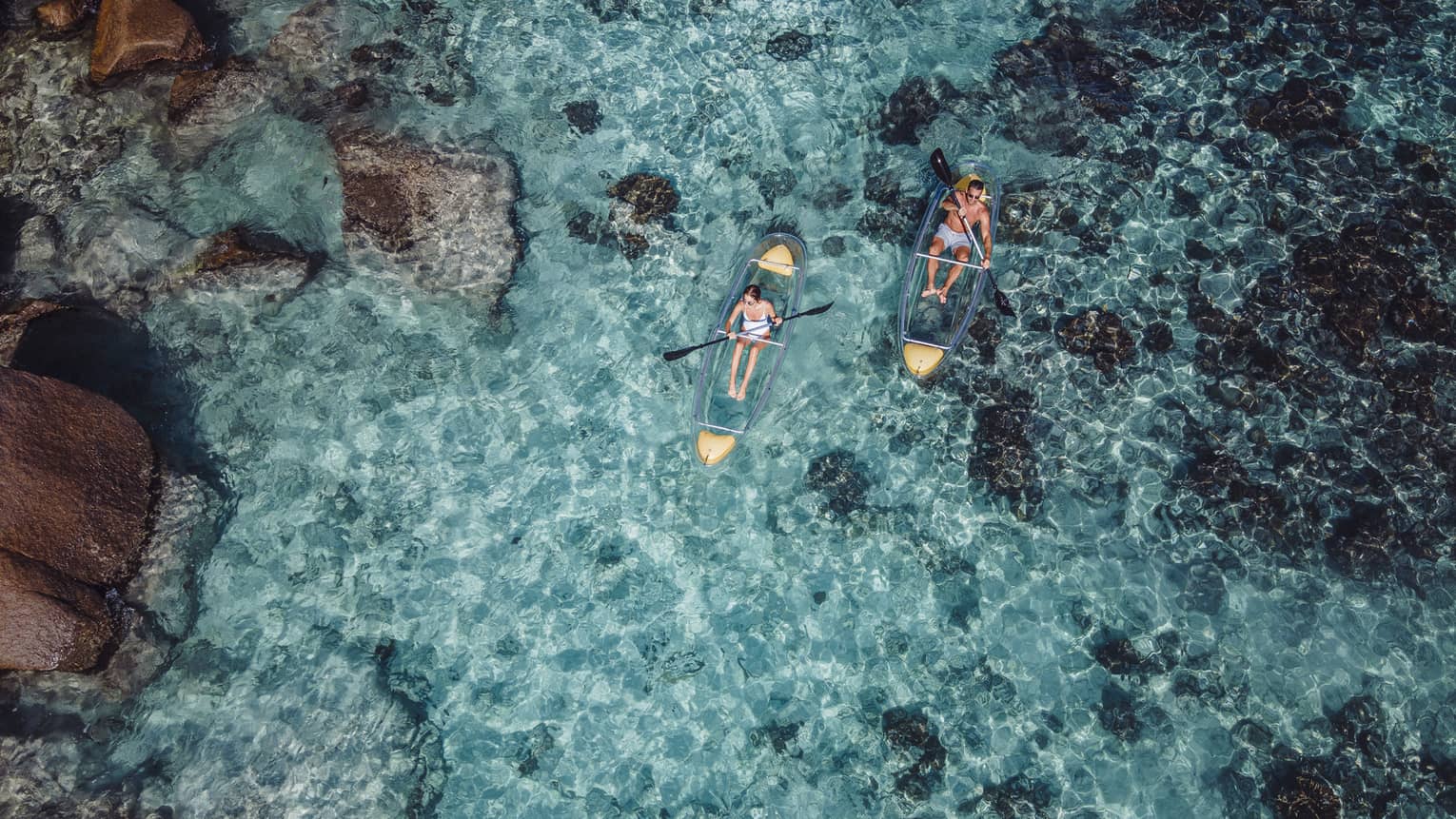 A view from above of kayakers paddling in transparent kayaks near boulders jetting out of crystal-clear open water.