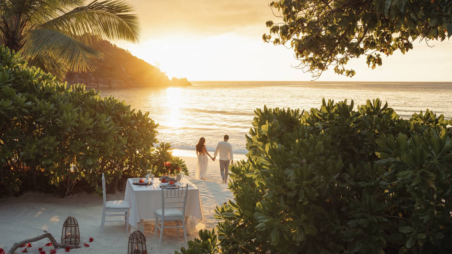 Framed by trees, a romantic dinner table set for two on the beach; a couple holding hands walks in the golden glow of sunset.