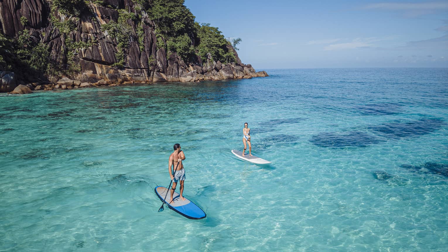 Two people stand-up paddleboarding on clear blue water with rocky cliff in back drop