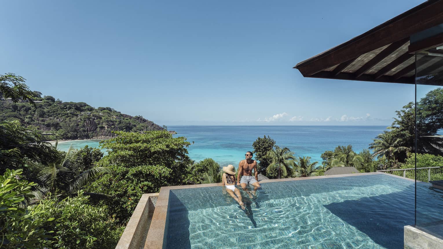 A couple lounges in a large infinity pool overlooking crystal blue waters
