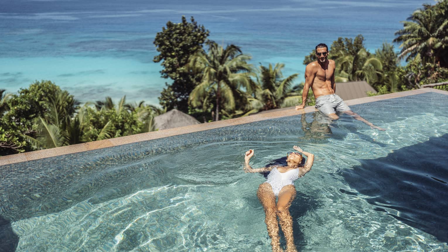 Two people in swimsuits, one floating on back in pool and one sitting on pool?s edge with palm trees and ocean in backdrop