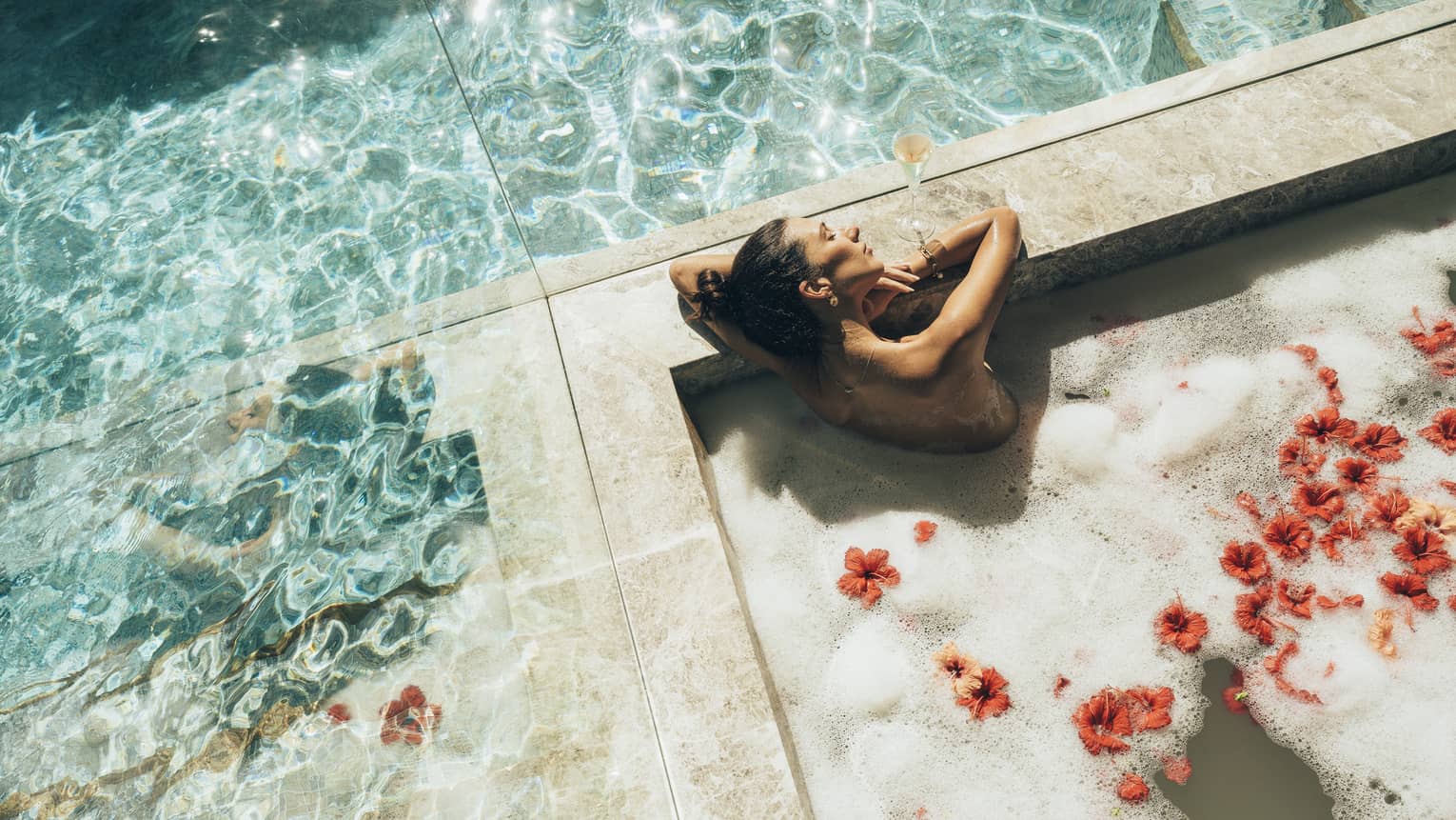 Surrounded by mirrored walls, a bather relaxes, eyes closed, in a marble tub of frothy water and red hibiscus blossoms.