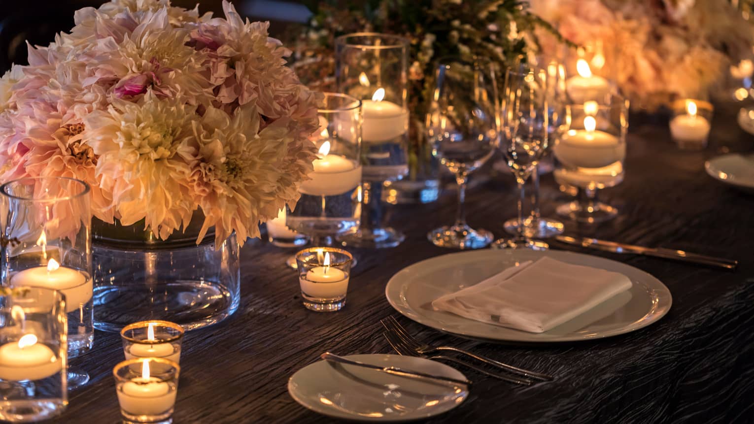 Pink and white flower arrangement in glass vase on dining table with glowing tealight candles