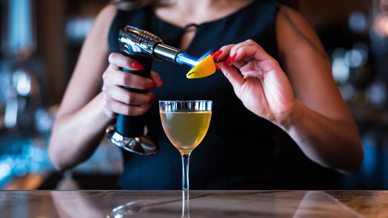 A woman using a blowtorch on an orange slide while a cocktail sits on the bar in front of her.