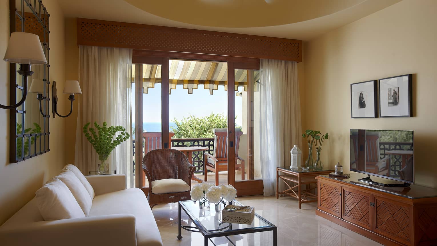 Two-Bedroom Sea-View Family Suite sunny living room with white sofa, wicker chair, open door to balcony with two chairs