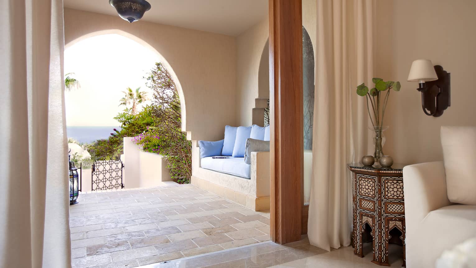 Covered balcony with cushions over clay sofa, tile floor, doorway to open-air room with decorative table, flowers