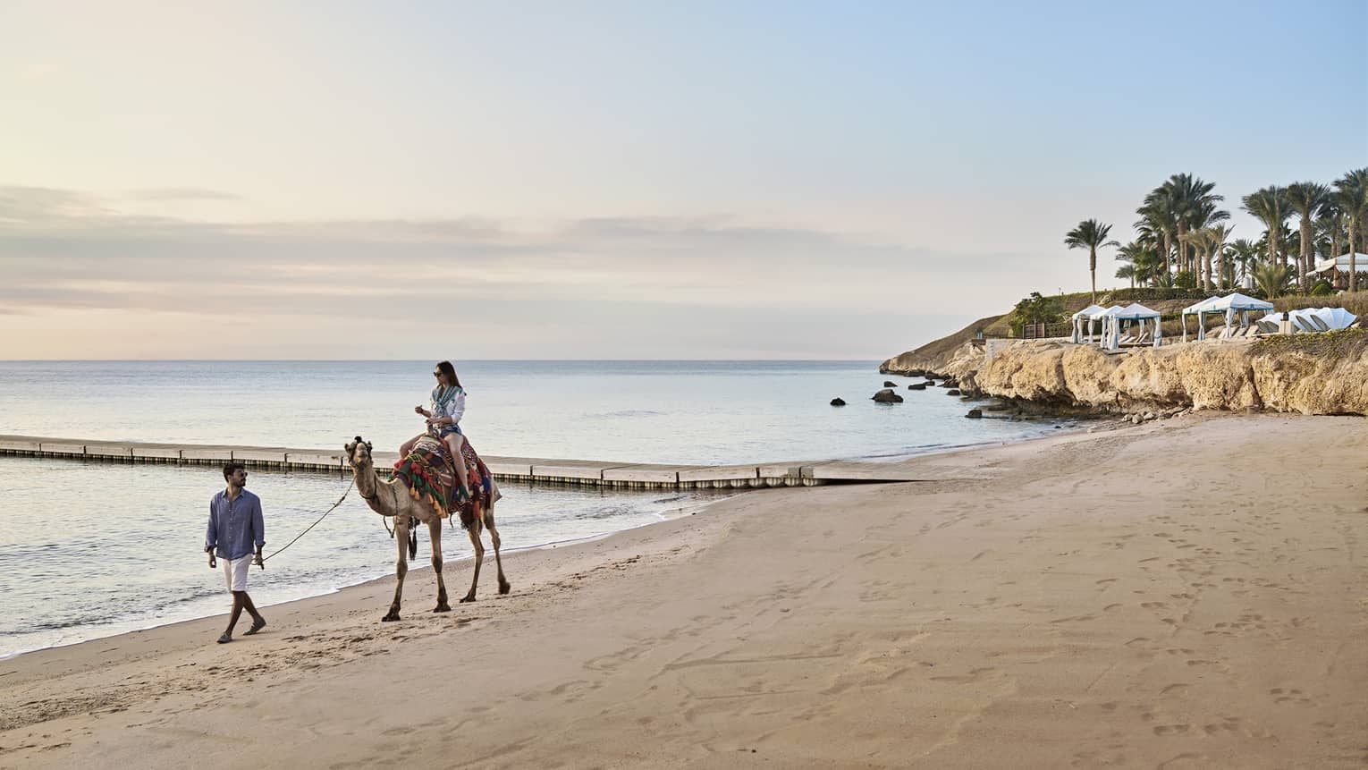 A woman rides a camel, led by a man, along the beach at Four Seasons Sharm El Sheikh