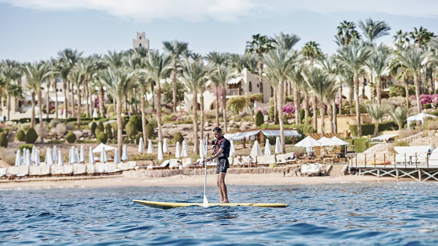 A paddler stands on a board in the water in front of a beach lined with lounge chairs and palm trees and a villa beyond.