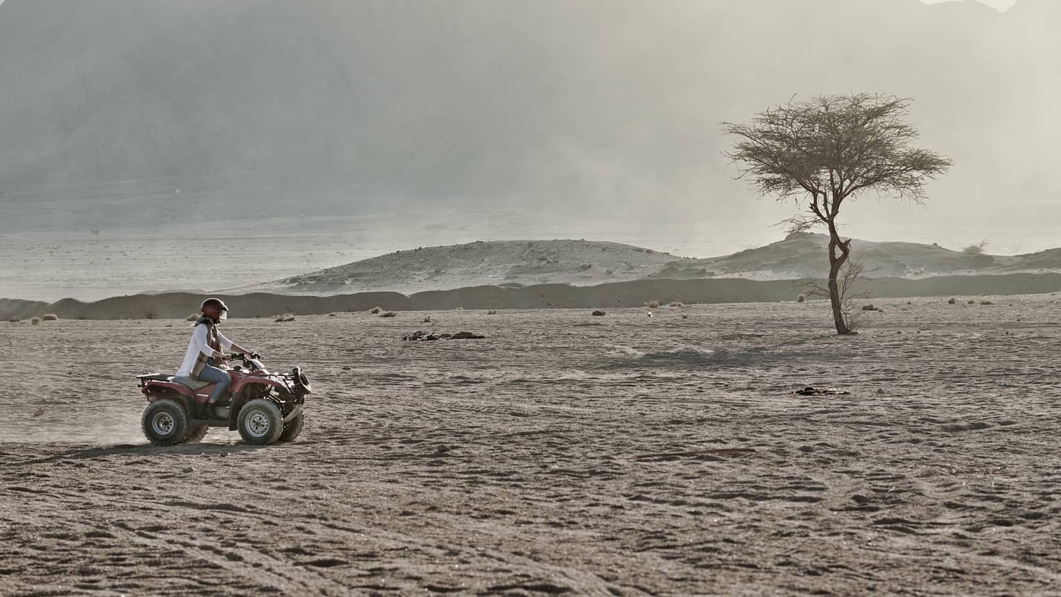A person in a helmet rides an ATV over an arid, dusty landscape past large hazy mountains that fill most of the image.