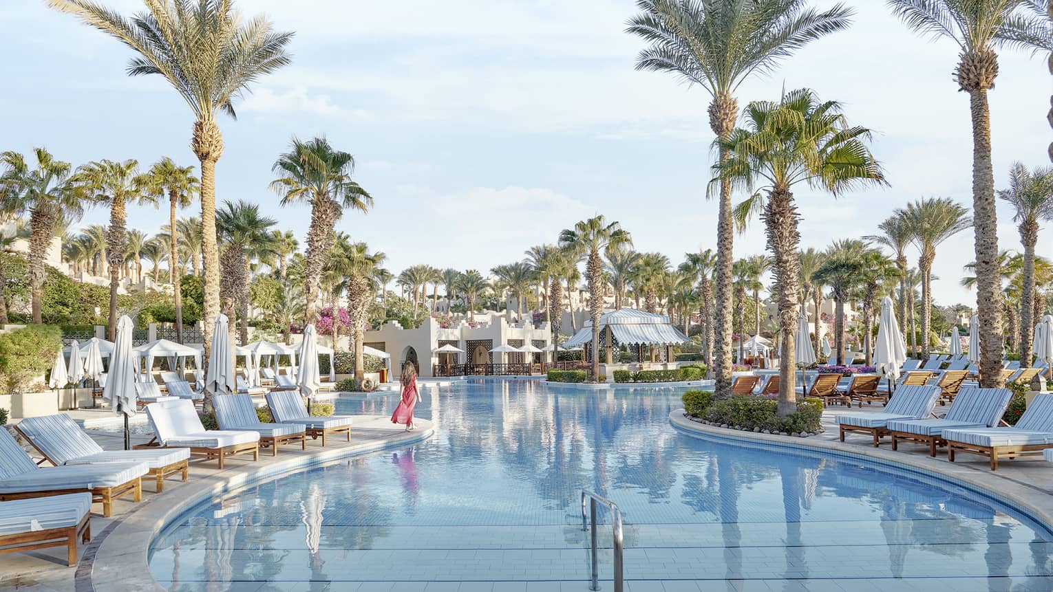 A woman walking along a pool surrounded by palm trees.