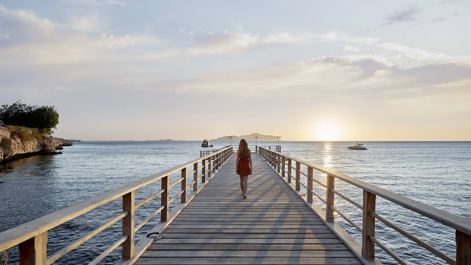 A woman walking along a long wooden walkway that extends into the ocean, the sun is setting in the background.