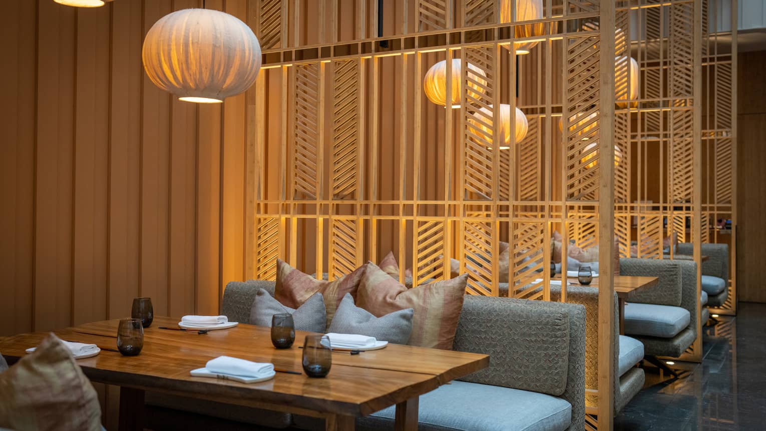 Restaurant with delicate wood slat partitions dividing plush banquette seating laden with pillows under golden lanterns.