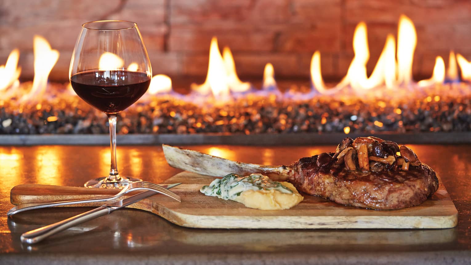 Glass of red wine, grilled chop on wood platter with cheese, mushrooms by fireplace