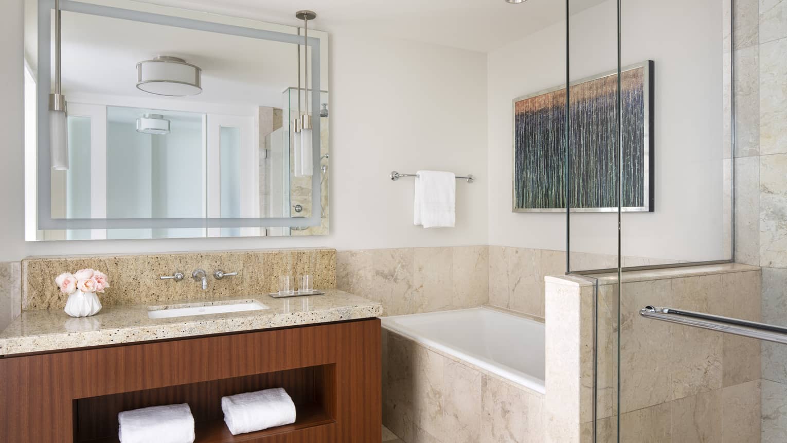 Luxury hotel bathroom with granite countertop vanity, mirror, glass-enclosed shower, a bathtub surrounded by beige tile, and wall art