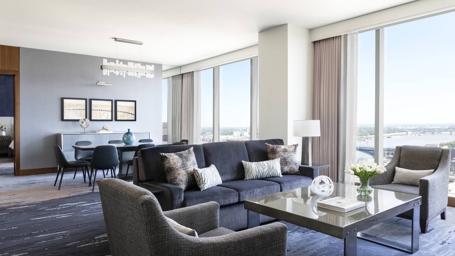 Hotel suite living and dining area with large windows offering a river view, dark gray sofa, armchairs, a coffee table and a dining table with modern chairs