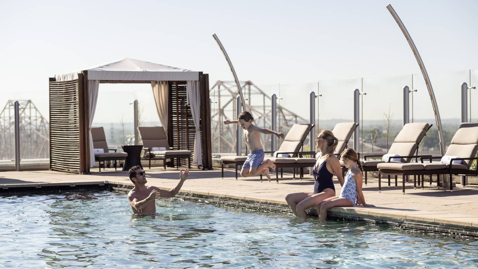 A family playing in an outdoor pool with lounge chairs and a cabana next to them.