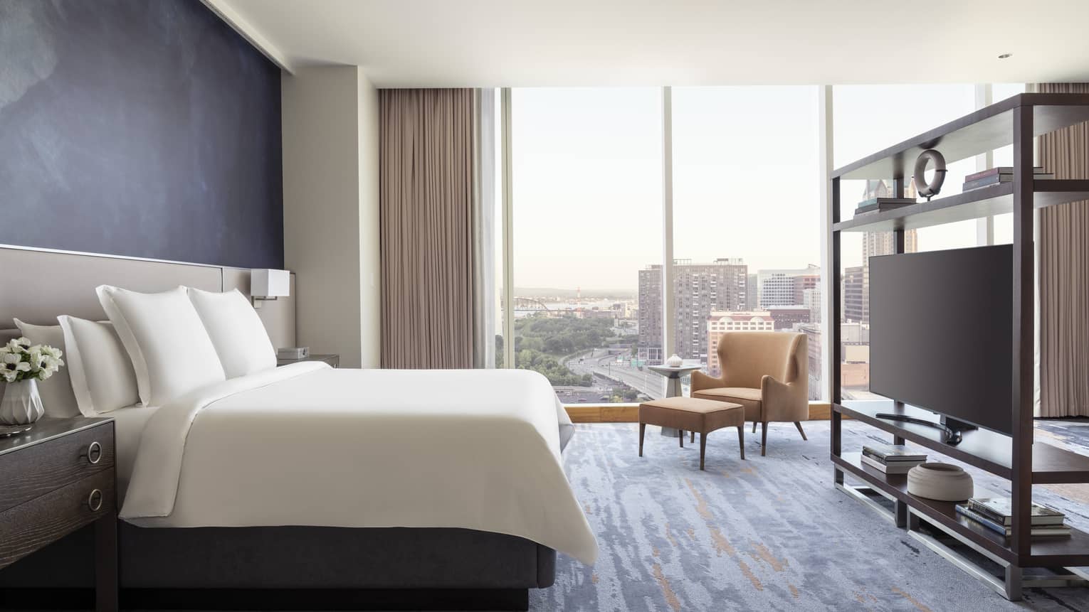 Luxury hotel room with a king bed, armchair and ottoman, shelving unit with a flat-screen TV, and floor-to-ceiling windows with a city view