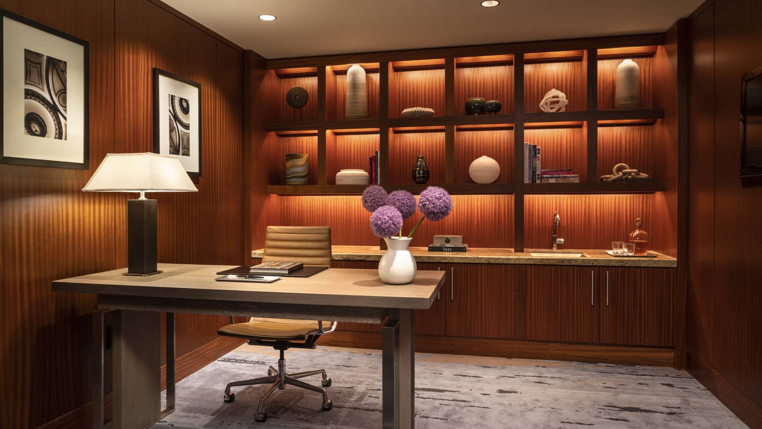 Luxury office room with wood-paneled walls, a desk, leather chair, backlit shelves with decorative items, and a vase of purple flowers