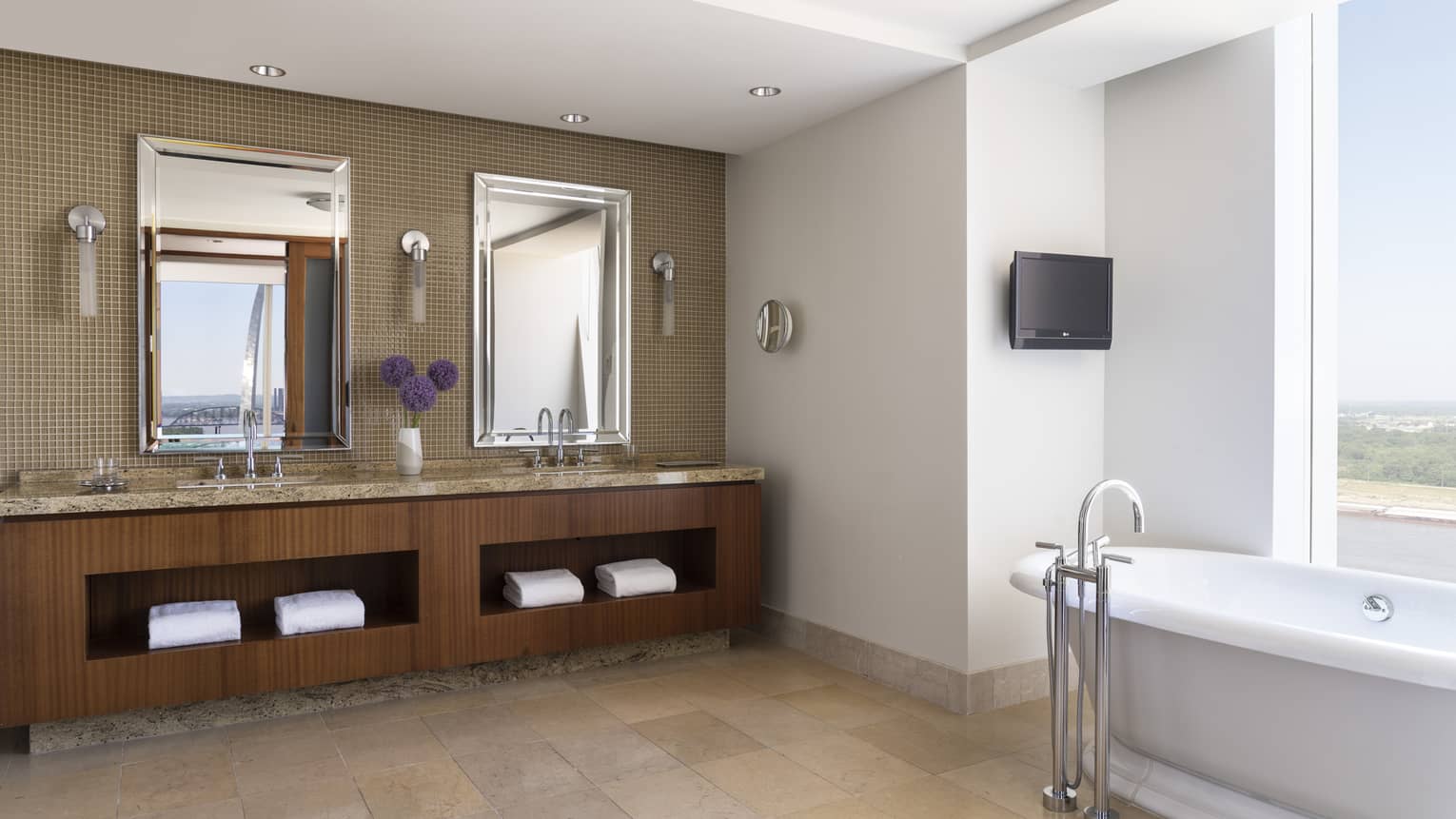 Luxury suite bathroom with a double vanity, mirrors, a freestanding bathtub by the window, and a wall-mounted TV